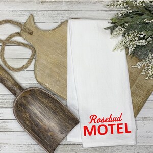 Op de afbeelding: Witte keukendoek met rode geborduurde tekst die "Rosebud Motel" zegt.