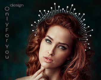 May include: A woman with long, curly red hair wears a silver crown of small, pointed spikes with white pearls at the ends. She is wearing a simple silver ring on her finger.