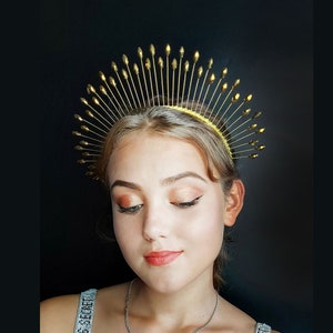 May include: A woman wearing a gold crown with a pointed design. She is wearing a silver necklace with a cross pendant and a white tank top with black text.