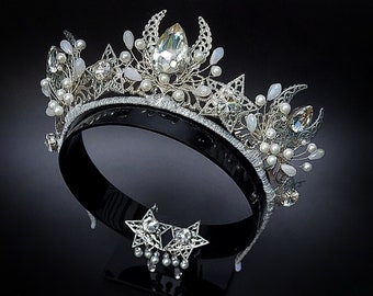 May include: A silver crown with a crescent moon design, adorned with white pearls and clear crystals. The crown is on a black stand with a matching star-shaped pendant.