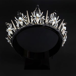 May include: A black crown with silver and white spider accents. The crown is made of fabric and has a black velvet band.