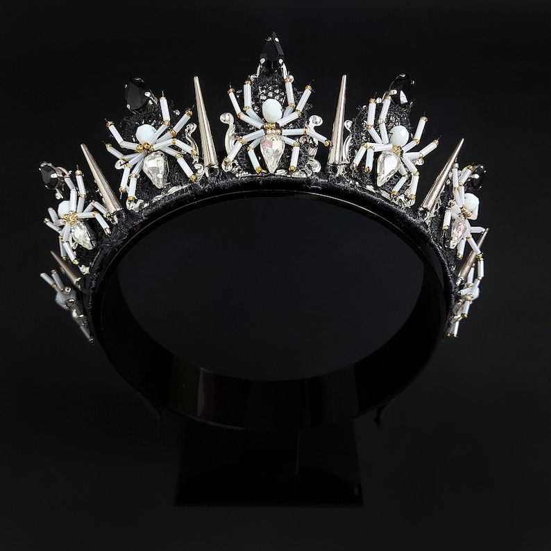 May include: A black crown with silver and white spider accents. The crown is made of fabric and has a black velvet band.