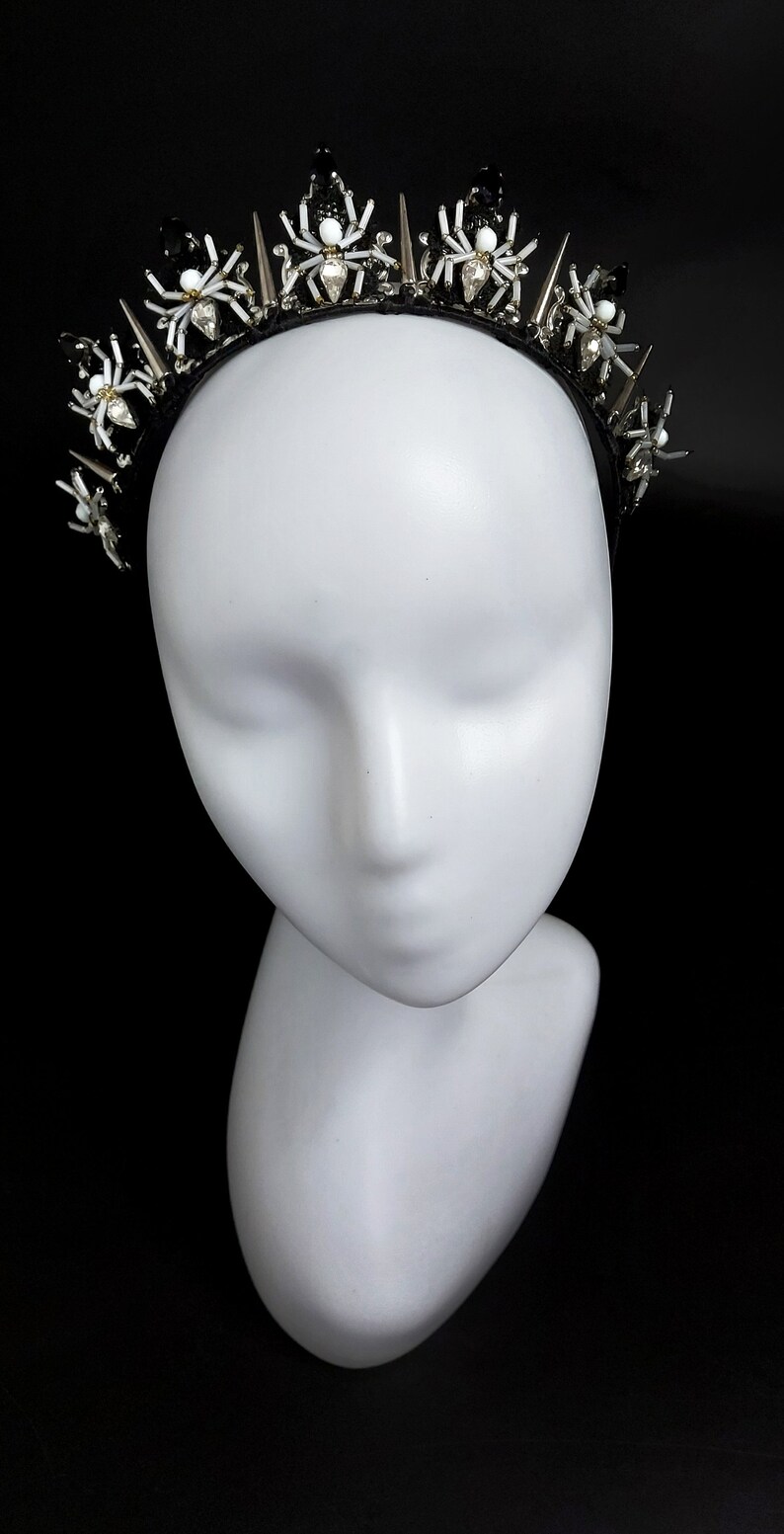May include: A black headband with silver spikes and white spider embellishments. The headband is on a white mannequin head.