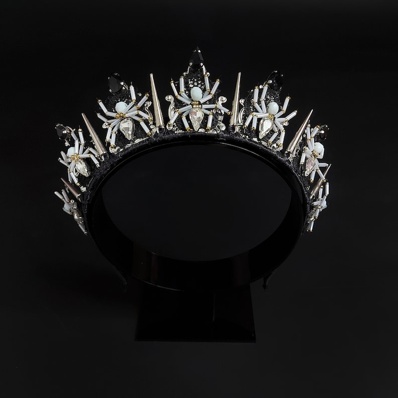 May include: Black crown with silver spikes and white spider embellishments. The crown is made of fabric and has a black velvet band.