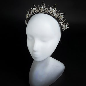 May include: A black headband with silver and white spider accents. The headband is decorated with small, sparkling rhinestones and has a pointed, crown-like shape.