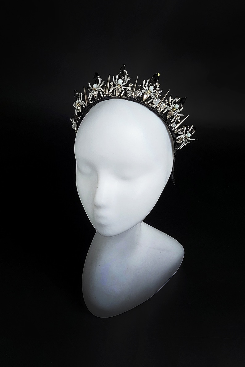May include: A black headband with silver and white spider accents. The headband is decorated with small, sparkling rhinestones and has a pointed, crown-like shape.