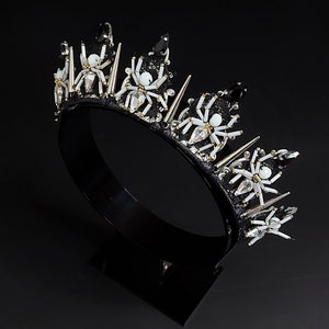 May include: A black crown with silver spikes and white spider embellishments. The crown is on a black stand.