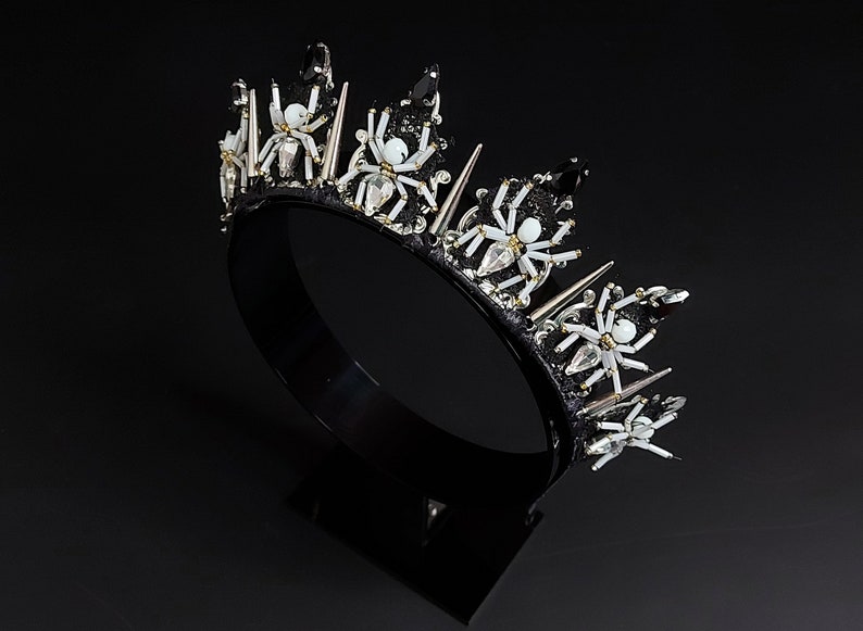 May include: A black crown with silver spikes and white spider embellishments. The crown is on a black stand.