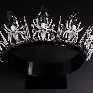 May include: A black crown with silver spikes and white spider embellishments. The crown is on a black stand.