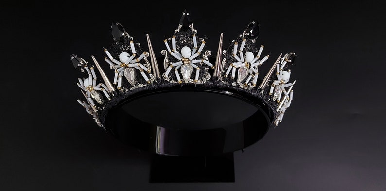 May include: A black crown with silver spikes and white spider embellishments. The crown is on a black stand.