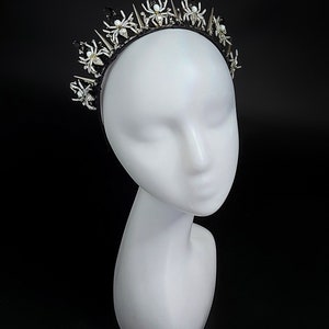 May include: A black headband with silver and white spider accents and spikes. The headband is worn on a white mannequin head.