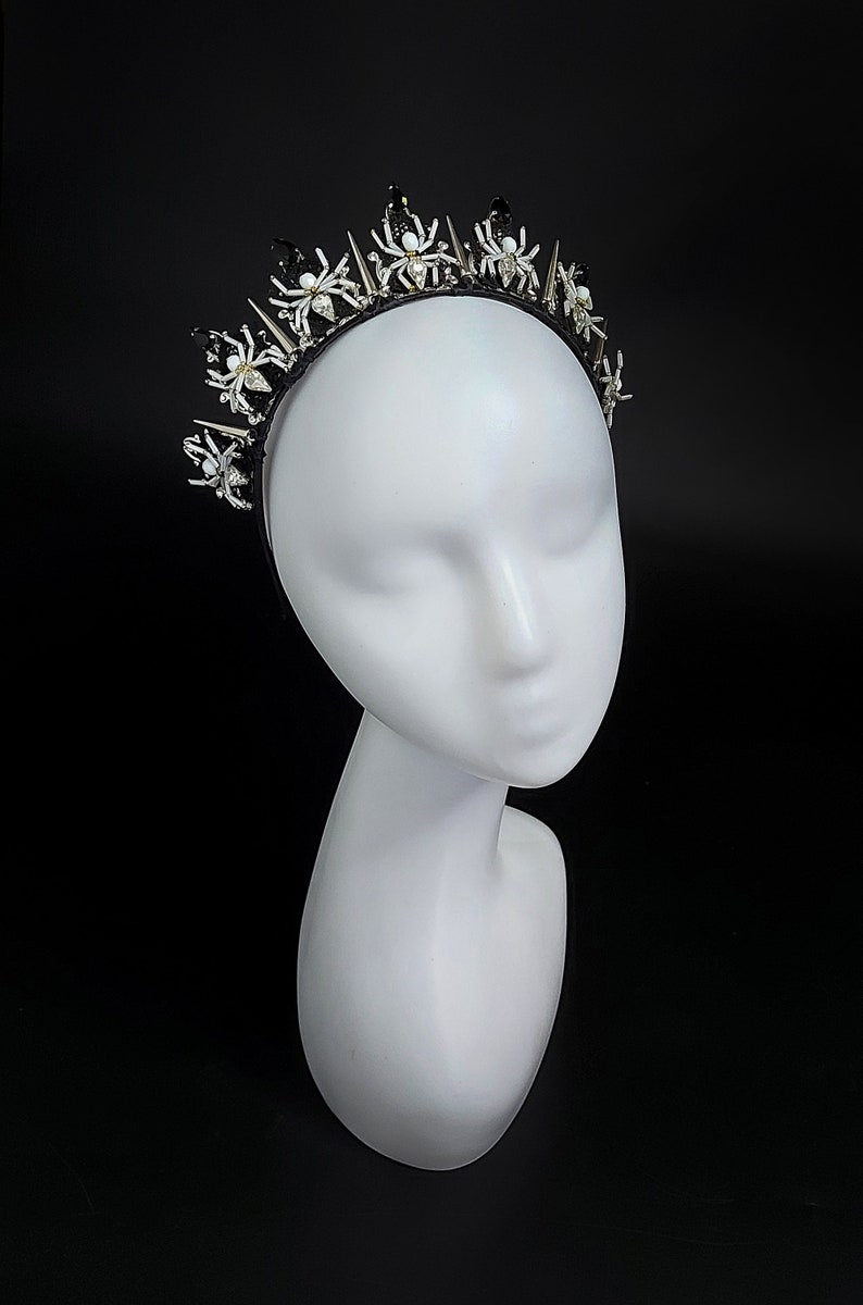 May include: A black headband with silver and white spider accents and spikes. The headband is worn on a white mannequin head.