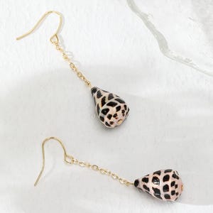 May include: A pair of gold dangle earrings featuring a black and white seashell pendant on each earring. The earrings are suspended from a gold chain.