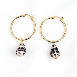 May include: A pair of gold hoop earrings with teardrop-shaped pendants. The pendants are white with black spots, resembling a seashell. The earrings are displayed on a white background.