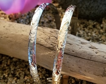 May include: Two silver-toned metal bangle bracelets featuring an intricate, embossed floral and scrollwork pattern across the entire outer surface. The bracelets have a polished, reflective finish and a rounded, slender profile, showcasing detailed engravings.