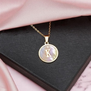 May include: A gold-colored necklace with a pendant featuring the letter "K" in a mother-of-pearl design. The pendant is circular and set in a gold frame. The necklace rests on a black box, with pink fabric in the background.