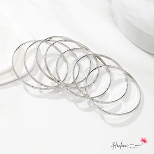 May include: A collection of seven silver bangles with a textured, patterned design. The bangles are arranged in a slightly overlapping manner, creating a layered effect. The jewelry is displayed on a white surface.