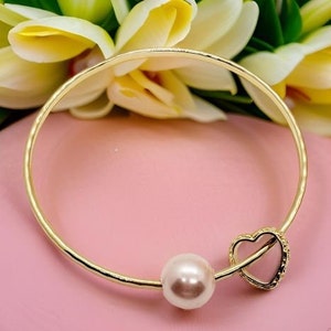 May include: A gold bangle bracelet with a heart charm and a pearl bead. The bracelet is on a pink surface.