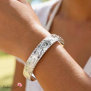 May include: A silver cuff bracelet featuring intricate engraved scrollwork and floral patterns across its polished surface. The metal band has a rounded profile with detailed etchings that catch the light, creating a reflective finish on the exterior.
