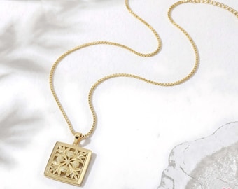 May include: Gold-toned necklace with a square pendant featuring a floral design. The pendant is intricately detailed with cut-out patterns. The chain is a delicate box chain style, and the necklace is displayed on a white surface.