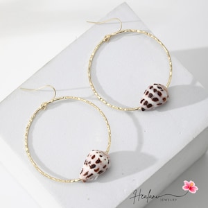 May include: A pair of gold-tone hoop earrings. Each earring features a textured hoop with a small seashell charm. The seashells are white with brown spots. The earrings are displayed on a white surface.
