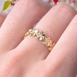 May include: A gold-toned ring featuring a continuous band of small, polished five-petaled flowers. The metal surface is smooth and reflective, creating a repeating floral pattern that encircles the finger with a bright, metallic finish.