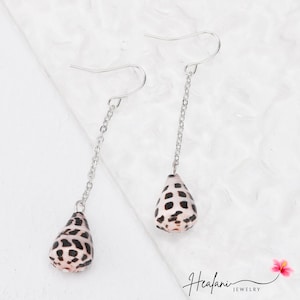 May include: Dangle earrings featuring teardrop-shaped shells with black spots, suspended from silver-tone chains and hooks. The shells have a natural, textured appearance. The earrings are displayed against a white background.