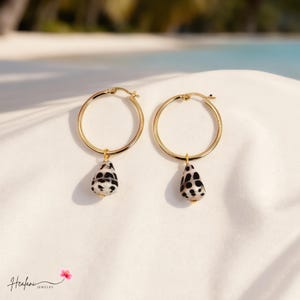 May include: A pair of gold hoop earrings, each featuring a dangling teardrop-shaped seashell charm. The shells are white with distinct black spots. The earrings include a hinged clasp mechanism at the top of the circular gold hoops.