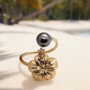 May include: A gold-toned open ring featuring a polished dark gray spherical pearl on one end and a detailed five-petaled hibiscus flower design on the other end. The metal band is smooth and circular, connecting the floral motif to the pearl.