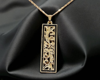 Op de afbeelding: Een goudkleurige rechthoekige hanger aan een ketting met een ingewikkeld opengewerkt bloemmotief tegen een zwarte achtergrond. De hanger hangt aan een bijpassende goudkleurige ketting met een gestructureerd schakelpatroon voor een geheel.
