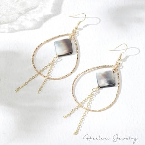 May include: A pair of gold-colored teardrop earrings. Each earring features a square, iridescent, gray-toned shell accent and dangling gold chain. The earrings have a textured finish and are displayed on a white surface. The brand name "Healani Jewelry" is visible.
