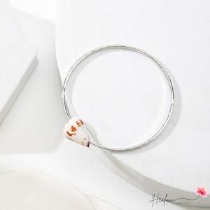 May include: A silver bangle bracelet with a seashell accent. The bracelet is a simple, thin band with a hammered texture. The seashell is white with brown and orange markings. The jewelry is displayed on a white surface.