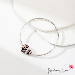 May include: A silver-colored bracelet featuring a small, natural seashell with a speckled pattern of brown and black. The seashell is attached to the bracelet's circular band. The bracelet is displayed on a white surface.