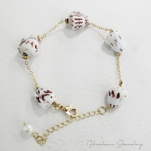 May include: A gold-toned chain bracelet featuring white seashells with brown markings. The bracelet has a lobster clasp closure and a small pearl at the end of the chain. The bracelet is labeled "Healani Jewelry".