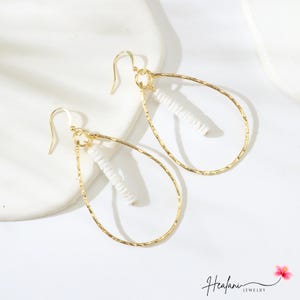 May include: A pair of gold-colored teardrop earrings with a hammered texture. Each earring features a white, cylindrical bead cluster suspended within the teardrop frame. The earrings have a hook closure. The jewelry is displayed on a white surface.