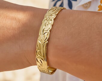 Op de afbeelding: Een goudkleurige armband met een ingegraveerd bloemen- en krulpatroon over het gehele oppervlak. Het metaal heeft een gepolijste, reflecterende afwerking met geschulpte randen die de contouren van het reliëfontwerp volgen.