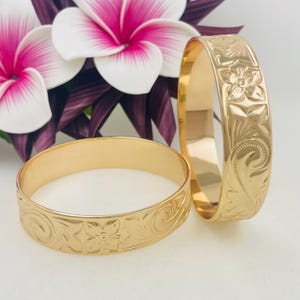 May include: Two gold-toned metal bangles featuring an engraved floral pattern with swirling leaf details. The bracelets have a polished, reflective surface and a smooth interior finish, showcasing a consistent design across the outer band.