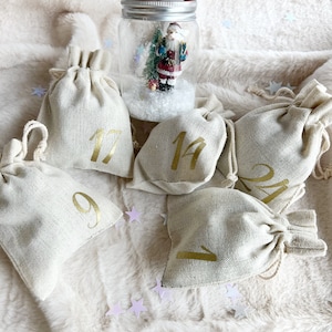 Pode incluir: Um conjunto de calendário do advento com pequenos sacos bege com cordão e números dourados, e um globo de neve com uma figura do Pai Natal. Os sacos estão dispostos sobre uma superfície branca e macia, com confetes em forma de estrela.