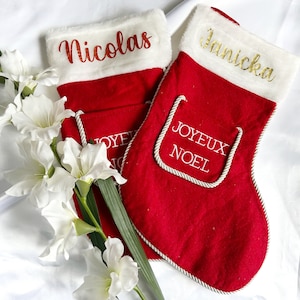 Könnte beinhalten: Zwei rote Weihnachtsstrümpfe mit weißen Kunstfellbündchen. Einer ist mit dem Namen "Nicolas" in kupferfarbener Schrift personalisiert, der andere mit "Janicka" in Gold. Die Worte "JOYEUX NOEL" sind auf der Vorderseite gestickt.