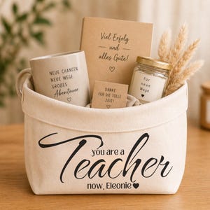 Könnte beinhalten: Ein cremefarbener Stoffkorb mit dem schwarzen Text "you are a Teacher now, Eleonie", der eine beige Tasse mit deutschem Text, ein braunes Notizbuch, ein kleines Glas mit goldenem Deckel und getrocknete Ziergräser enthält.