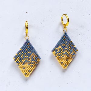 May include: A pair of diamond-shaped beaded earrings featuring a geometric pattern of metallic gold and muted periwinkle blue glass beads. Each earring hangs from a polished gold-tone metal hoop closure, creating a cohesive set of jewelry.