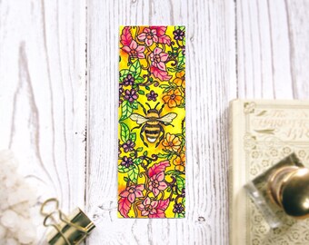 May include: A yellow bookmark with a bee and floral design. The bee is in the center of the bookmark and is surrounded by pink, yellow, and green flowers.