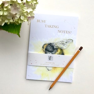 May include: A white notebook with a bee illustration and the text "Busy Taking Notes?" on the front cover. The notebook is 100% recycled and has 80 plain pages. The notebook is sitting on a white surface with a pencil next to it.