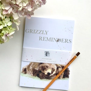 May include: A white notebook with the text "Grizzly Reminders" on the front cover. The notebook features a watercolour illustration of a brown bear sleeping on the front cover. The notebook is 100% recycled and has 80 plain pages.