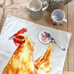 May include: A white tea towel featuring a watercolour illustration of a yellow chicken. The chicken is facing the left side of the image. The towel is on a wooden table with a white teapot, a white creamer, and a white mug with a chicken design.