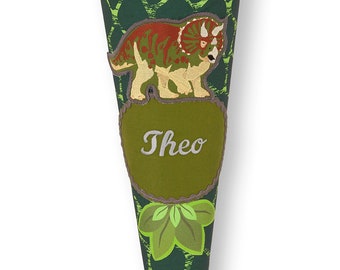 May include: Green and brown school cone with a dinosaur design and the name "Theo" embroidered on it. The cone is made of fabric and has a green and brown pattern. The cone is tied with a gray ribbon.