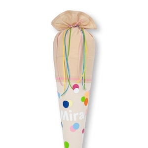 Könnte beinhalten: Ein beiger Stoffkegel mit einem rosa-weißen Punkte-Muster. Der Kegel hat ein weißes Namensschild mit der Aufschrift "Mira" und ist mit bunten Bändern gebunden.