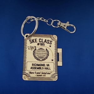 May include: A wooden keychain with a brown background and a silver key ring. The keychain has the text "SKE CLASS #185" and "RICHMOND, VA ASSEMBLY HALL" along with a small airplane and globe graphic. The text "Here I am! Send me!" and "Isaiah 6:8" is also included.