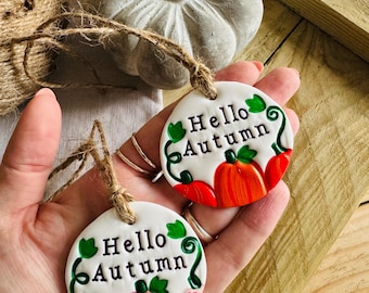 Può includere: Due ornamenti in ceramica bianca con il testo "Hello Autumn" e una zucca dipinta. L'ornamento superiore ha una zucca arancione e l'ornamento inferiore ha una zucca rosa.