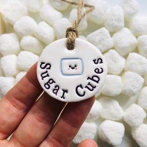 Op de afbeelding: Een witte keramische tag met de woorden "Sugar Cubes" in paarse letters. De tag heeft een illustratie van een lachende suikerklontje en een lus van jute touw om op te hangen. De achtergrond is gevuld met witte suikerklontjes.